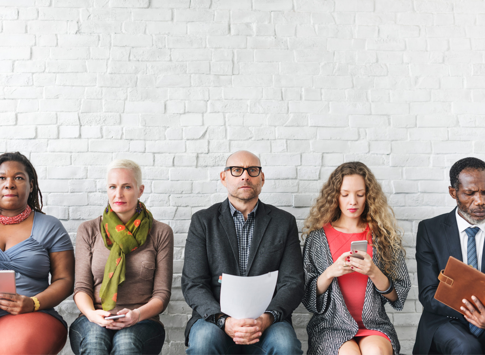 Five people sit in a row against a white brick wall, each focused on their phones or papers, displaying varied expres…