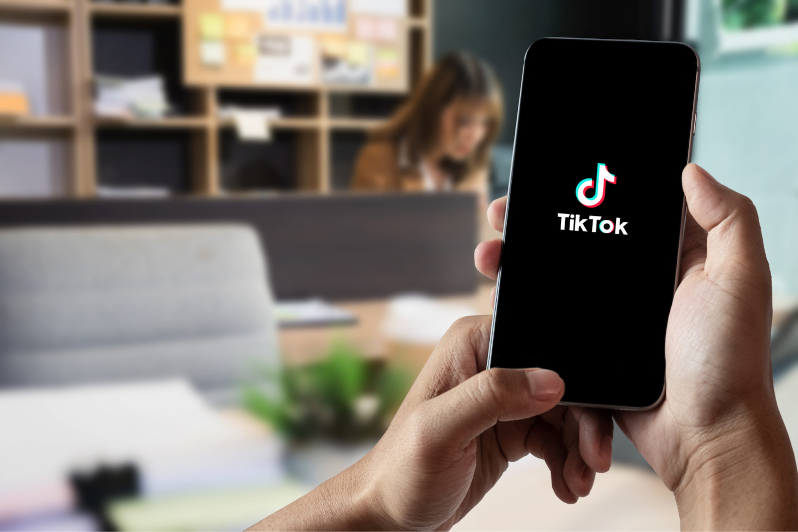 A person holds a smartphone displaying the TikTok logo, with a blurred office background